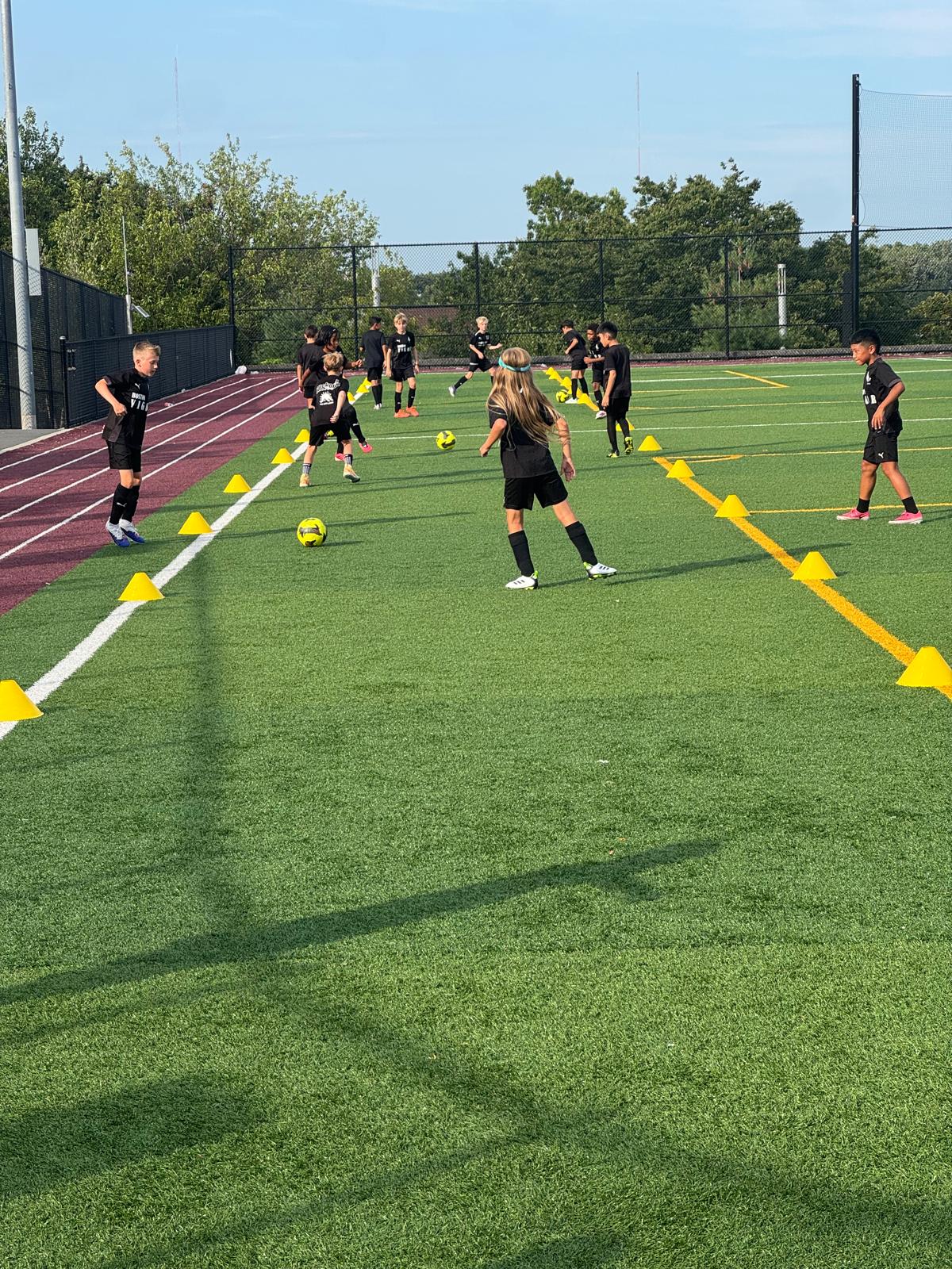 Young Boston Vigor FC players practicing dribbling drills around yellow cones on a turf field