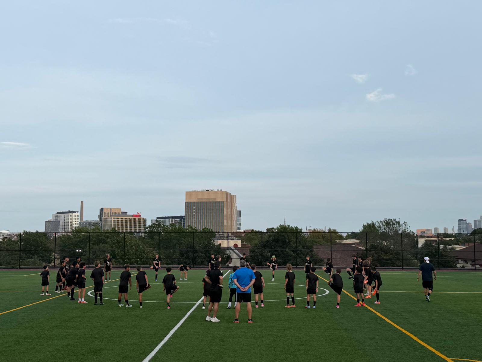Boston Vigor FC players training in a circle on a turf field with the Boston skyline behind them
